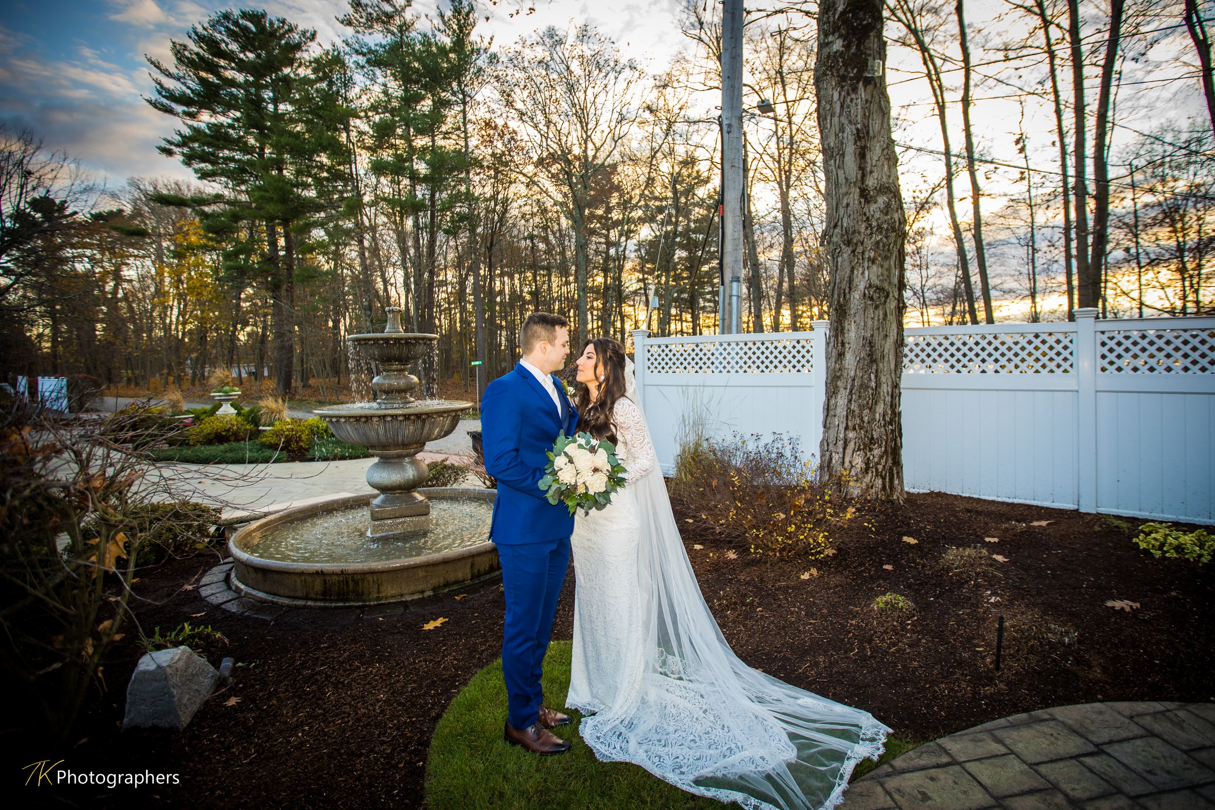 Bride and groom share a romantic moment by a garden fountain at sunset, surrounded by trees and white fence.