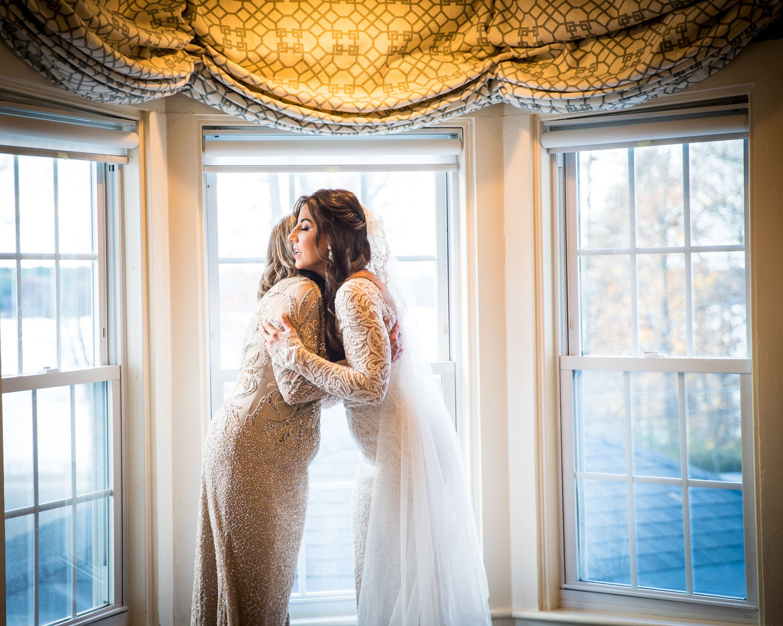 Bride and mother share an emotional hug in front of sunlit windows on wedding day.