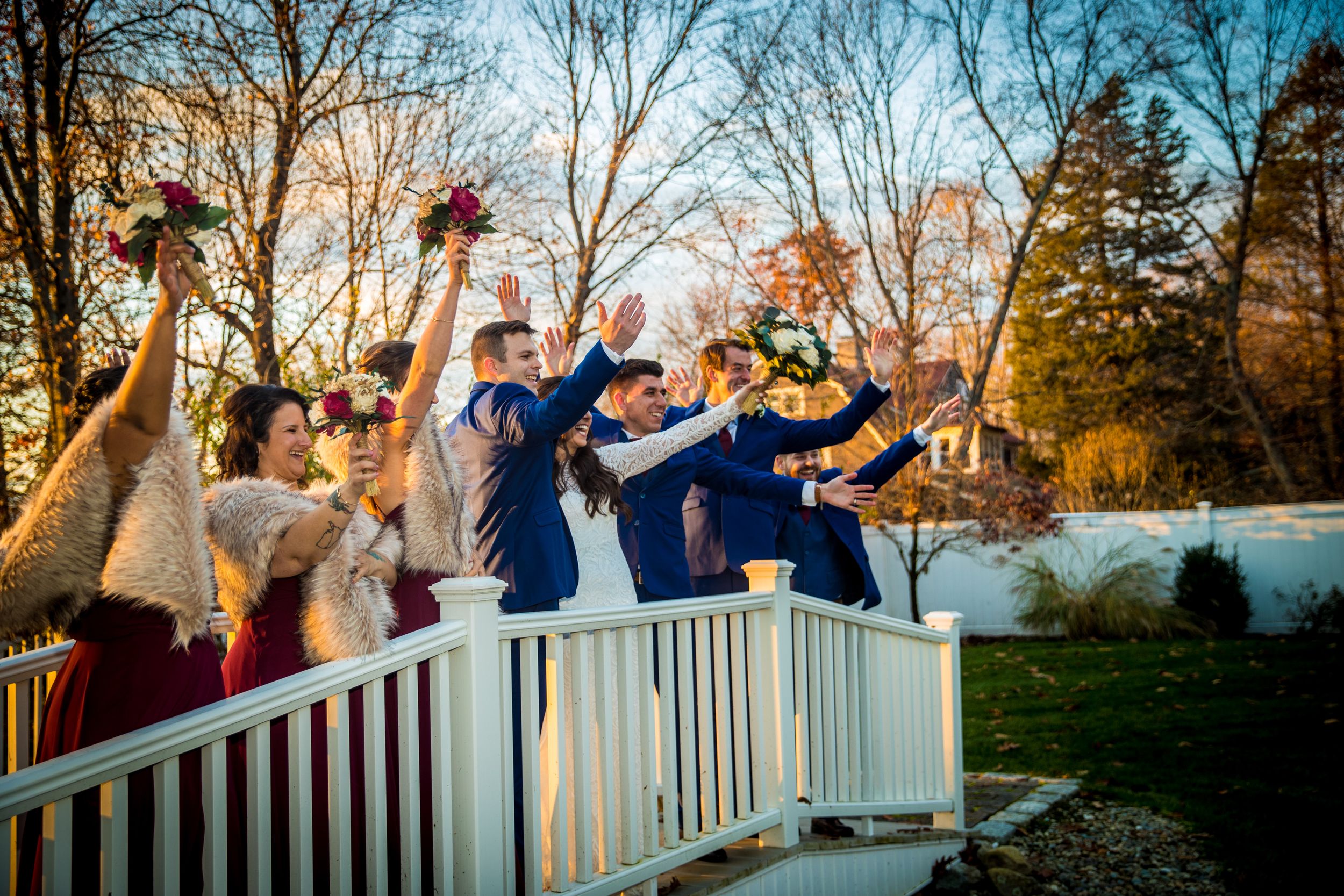 Wedding party celebrating on a white bridge, raising bouquets and hands in joy, surrounded by autumn trees at golden hour.
