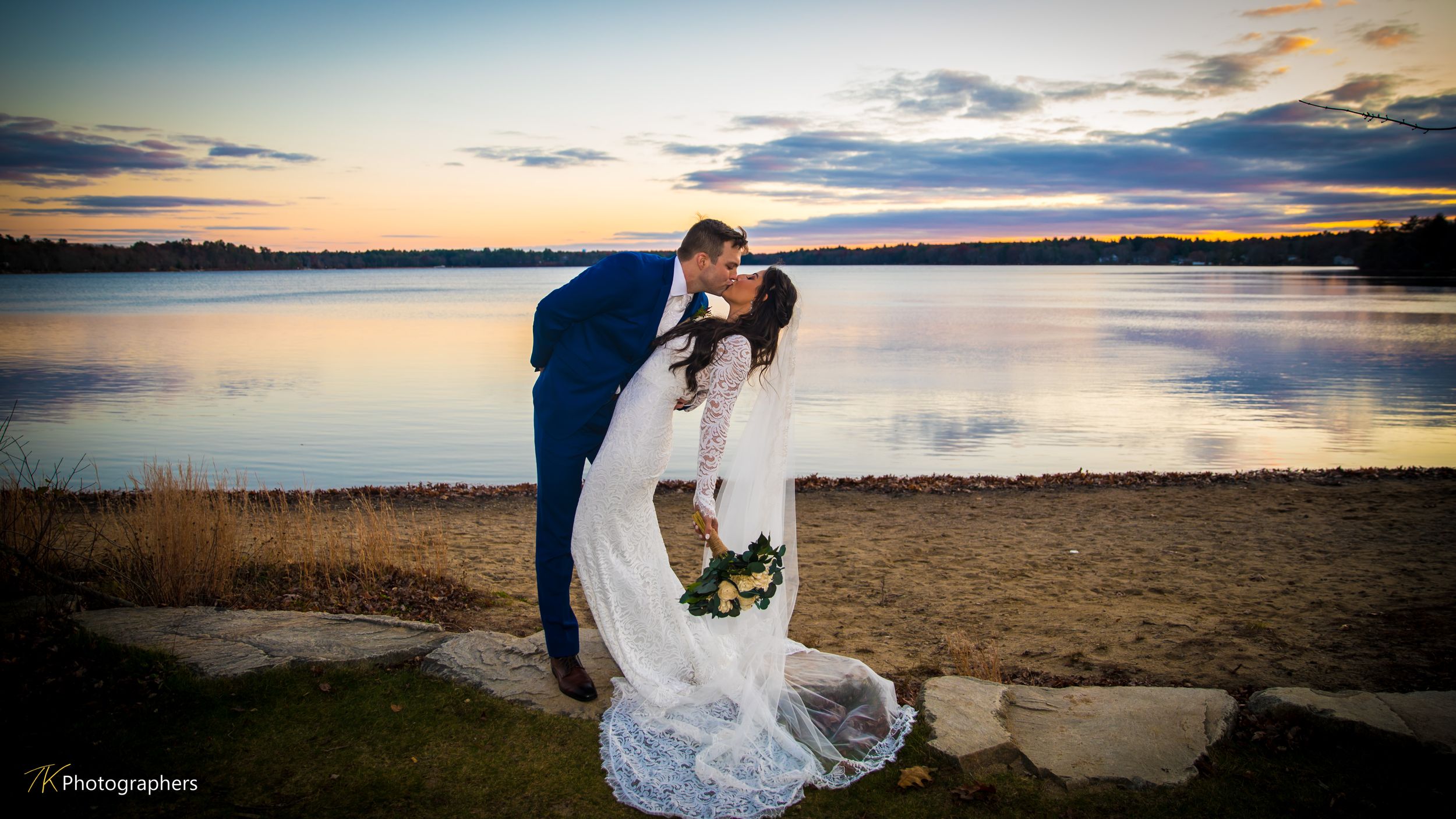 Bride and groom share romantic kiss at sunset by a serene lakeside, bride in white gown holding bouquet.
