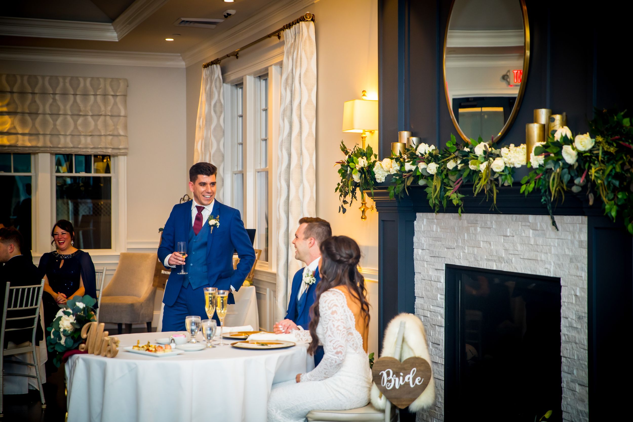 Best man gives toast to seated bride and groom at sweetheart table beside elegant fireplace with floral garland.