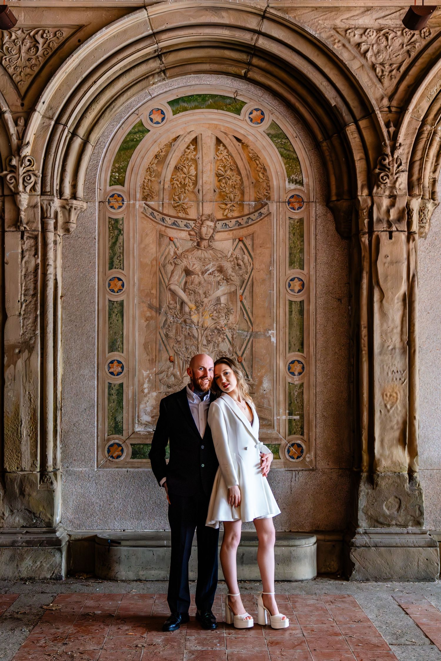Couple posing at Bethesda Terrace arcade in Central Park, NYC, with ornate stone carvings.