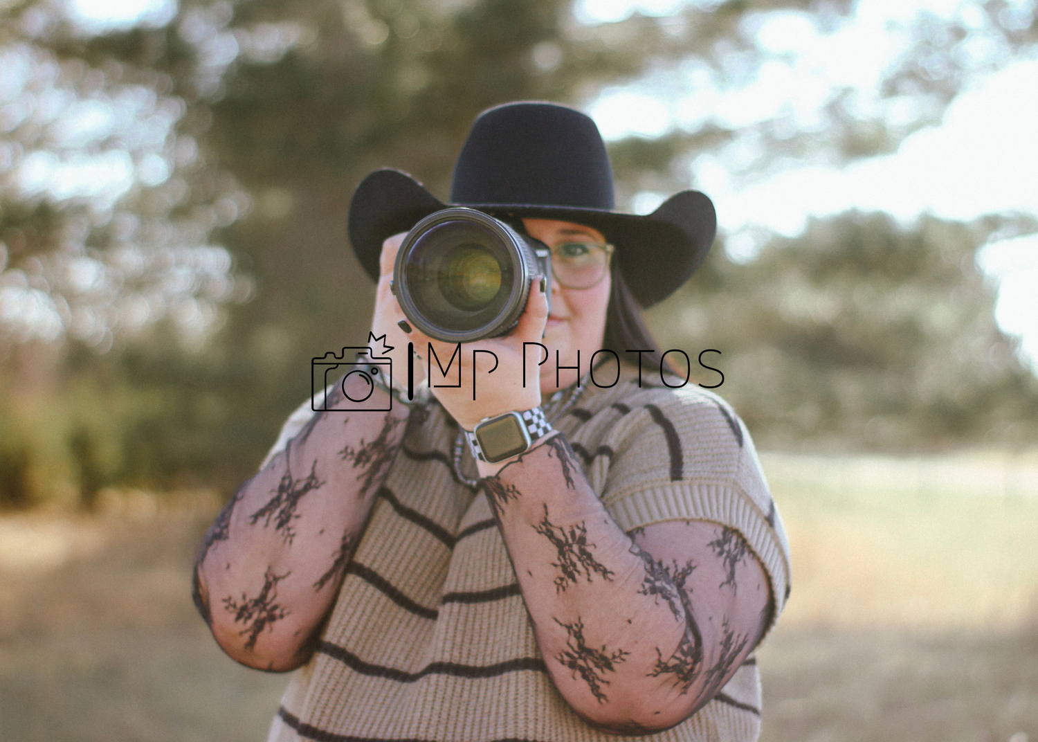 Tattooed photographer in black cowboy hat aims large camera lens toward viewer outdoors.