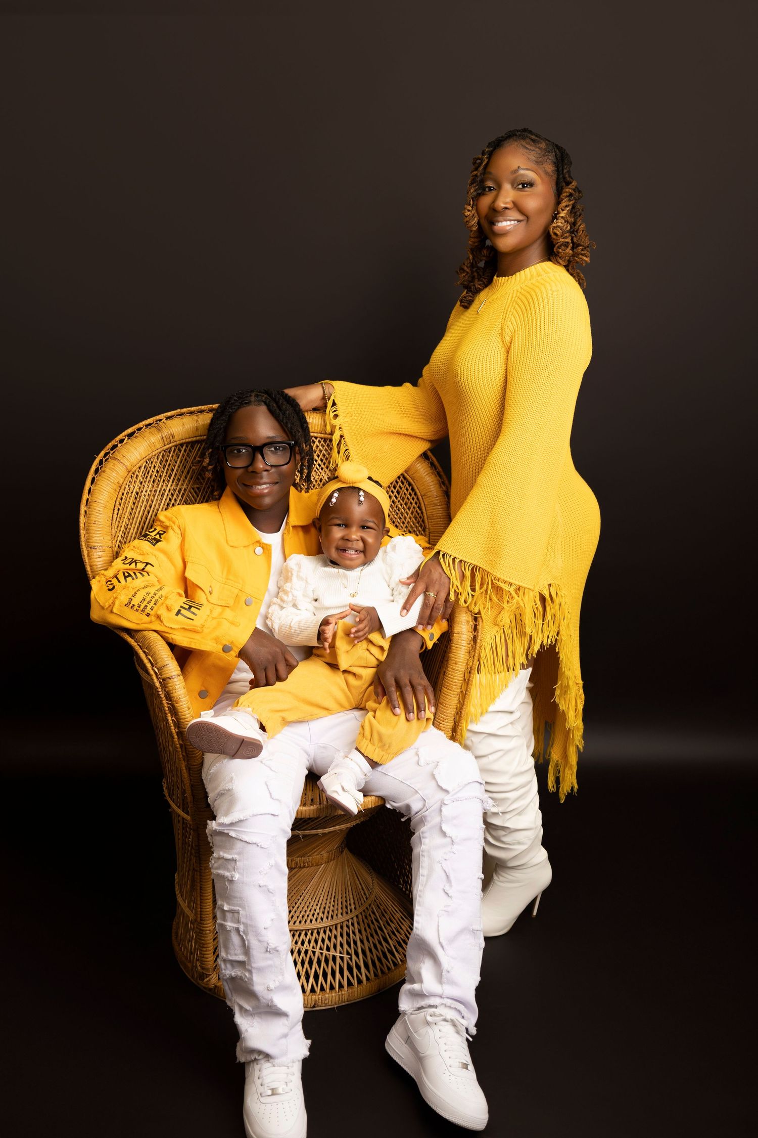 Family of three in matching yellow and white outfits posing in a wicker chair against a dark background.