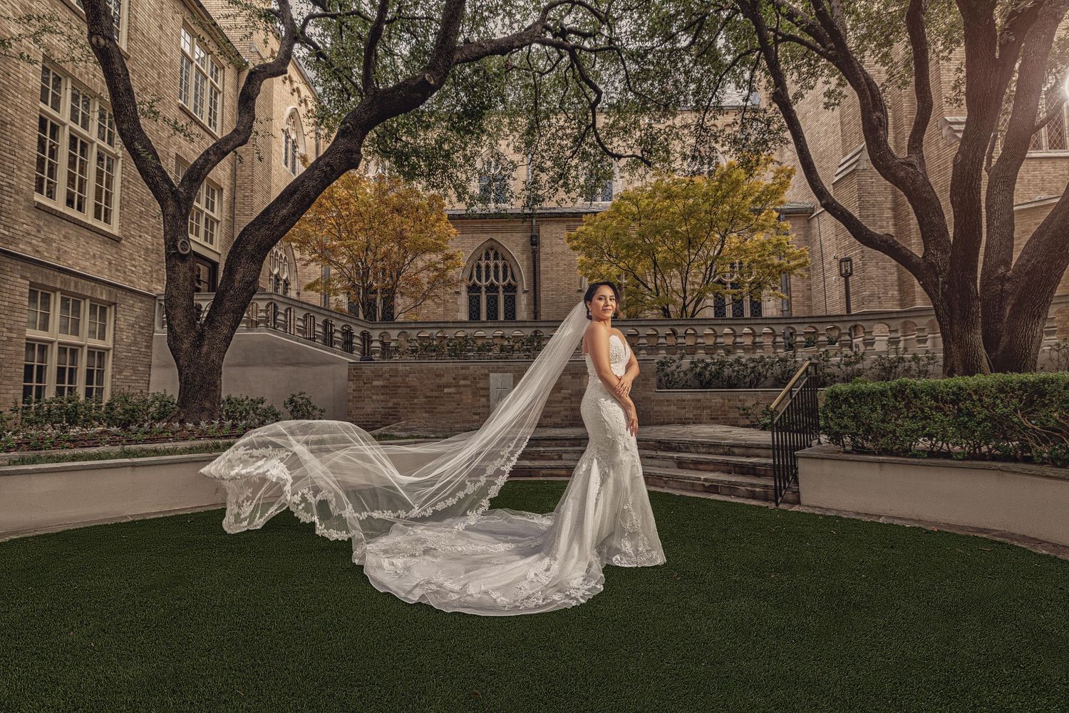 A bride in a lace mermaid gown poses in a stone church courtyard framed by mature trees, with her long train and veil flowing behind her