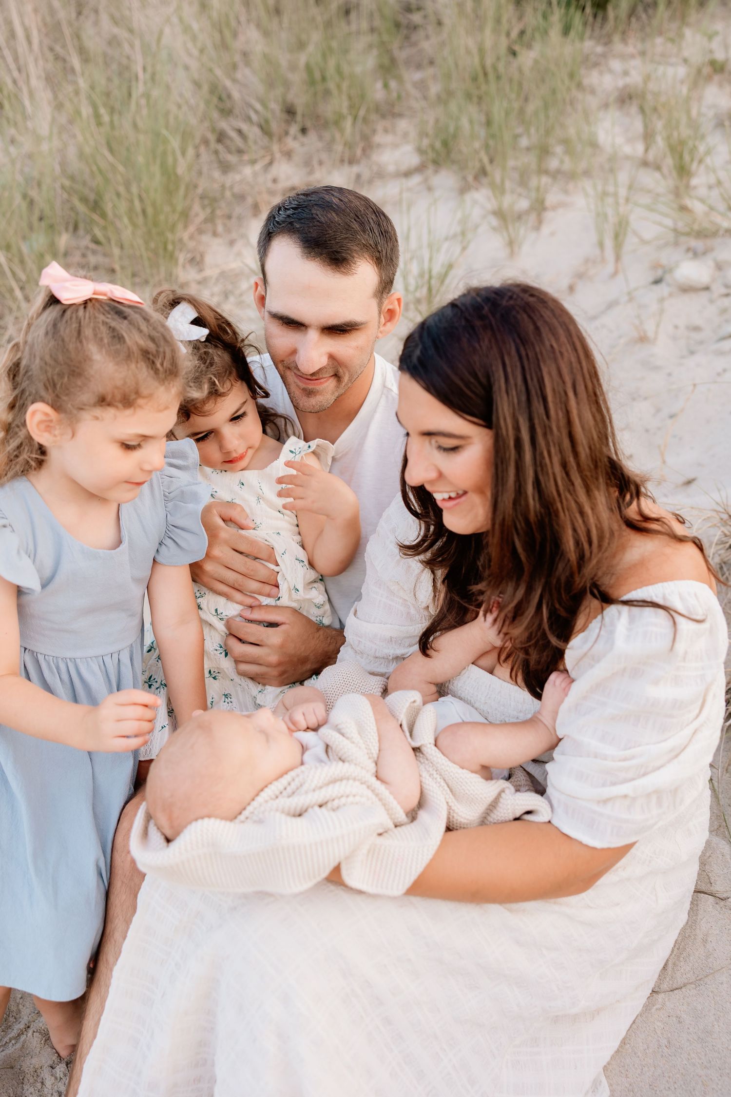 A happy family of five admires their newborn baby on a sandy beach, with two young girls in blue dresses.