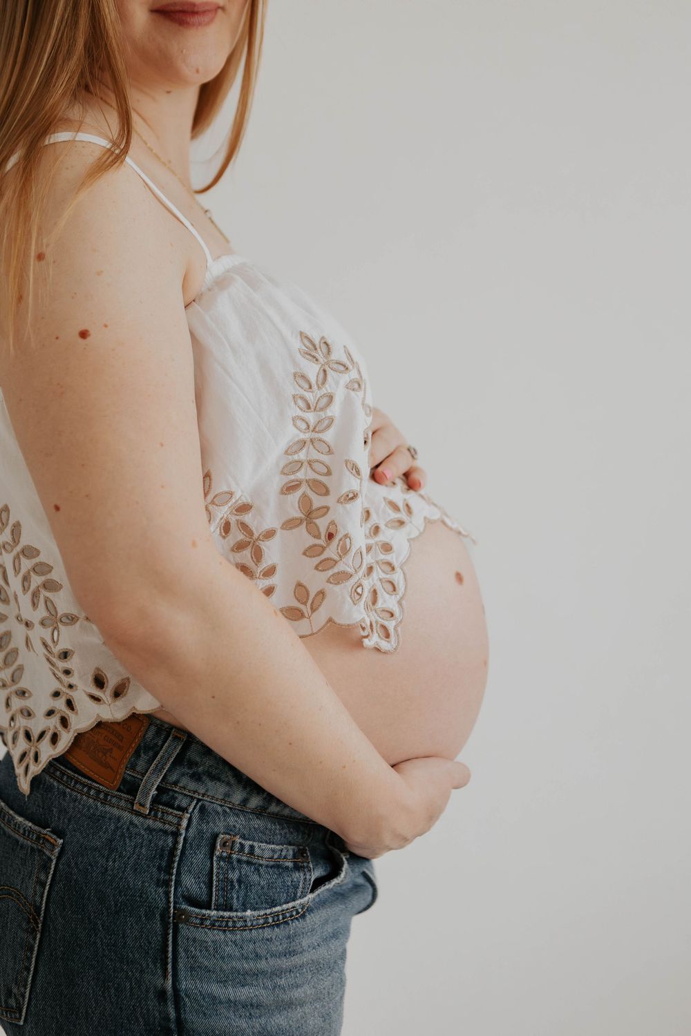 Close-up of pregnant woman in white lace top and jeans cradling bare baby bump. Cornwall Maternity Photographer