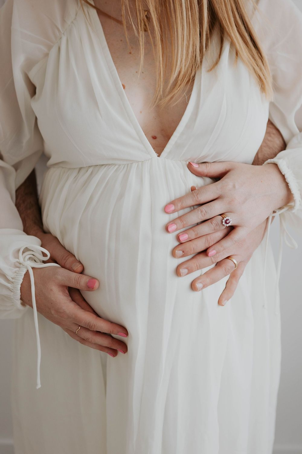 Couple tenderly holding pregnant belly in white maternity dress with partner hands visible. Cornwall Maternity Photographer