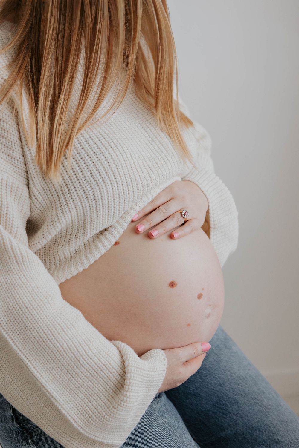 Pregnant woman in cream knit sweater and jeans gently holding bare baby bump indoors. Cornwall Maternity Photographer