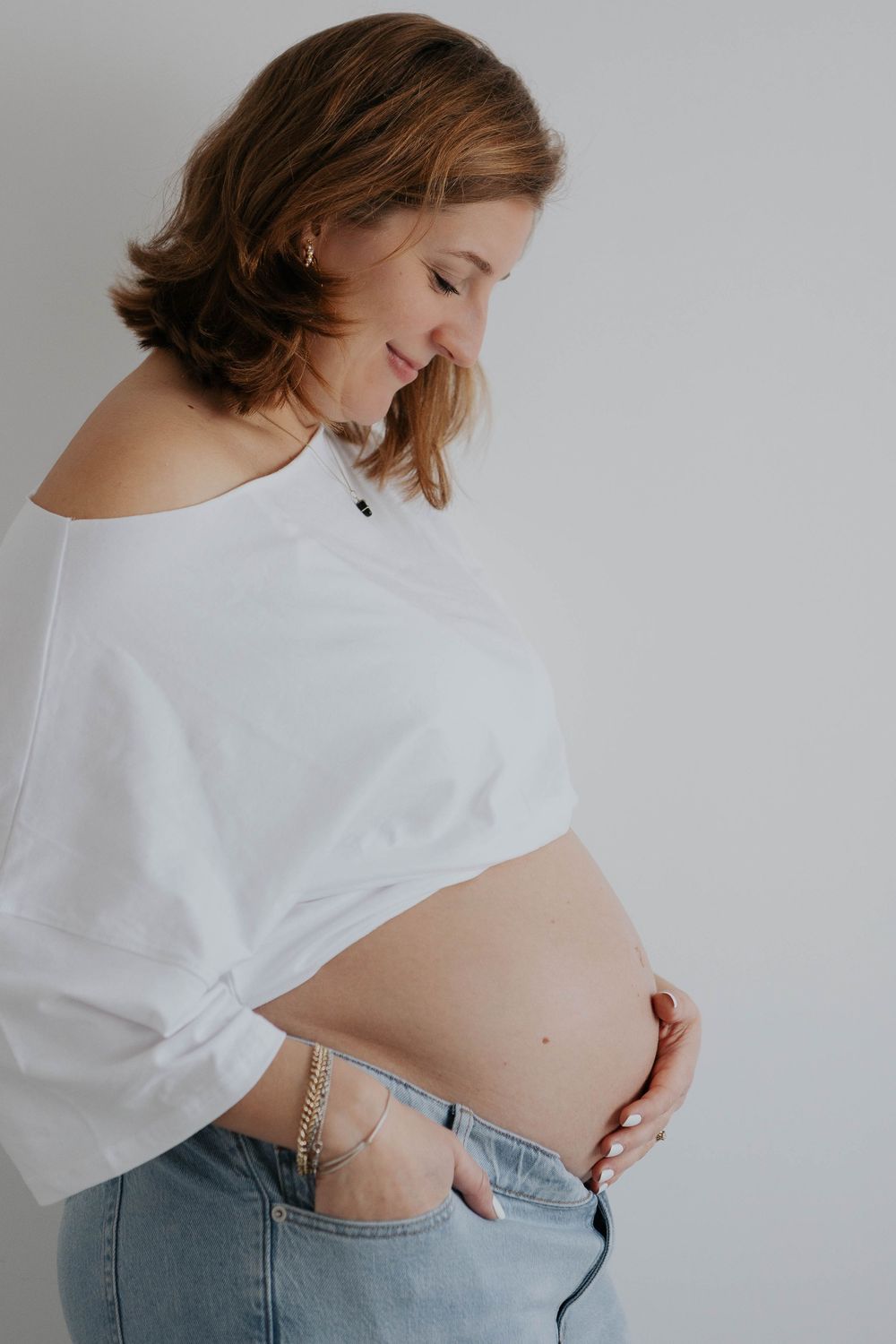 Smiling pregnant woman in white off-shoulder top and jeans cradling bare baby bump Cornwall Maternity Photographer