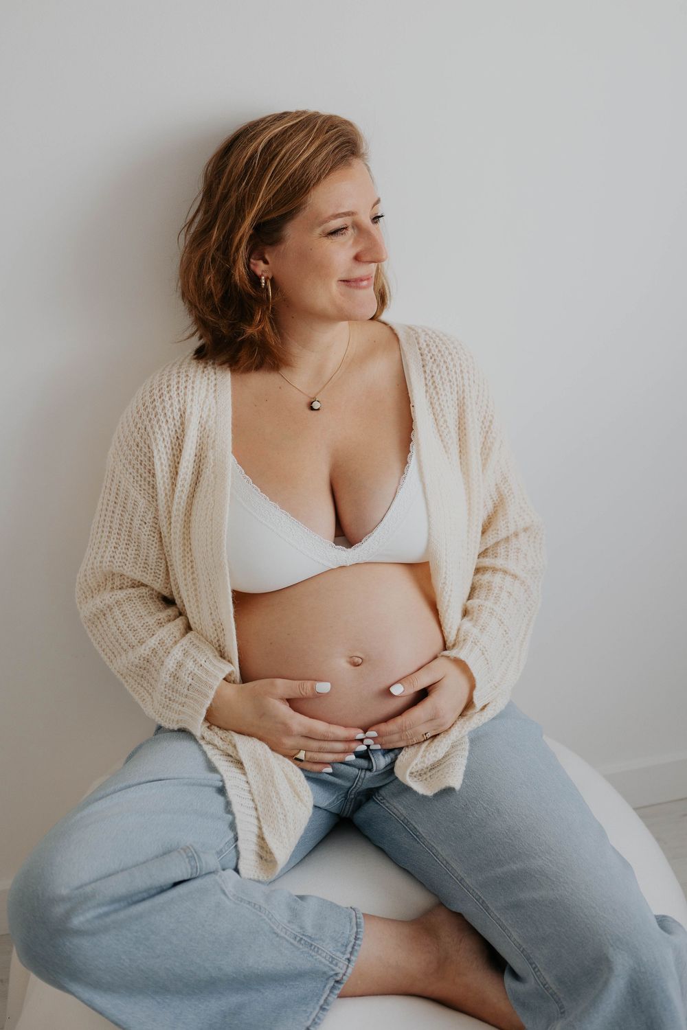 Pregnant woman in white bralette, cardigan, and jeans sitting and cradling her baby bump, smiling softly Cornwall Maternity Studio