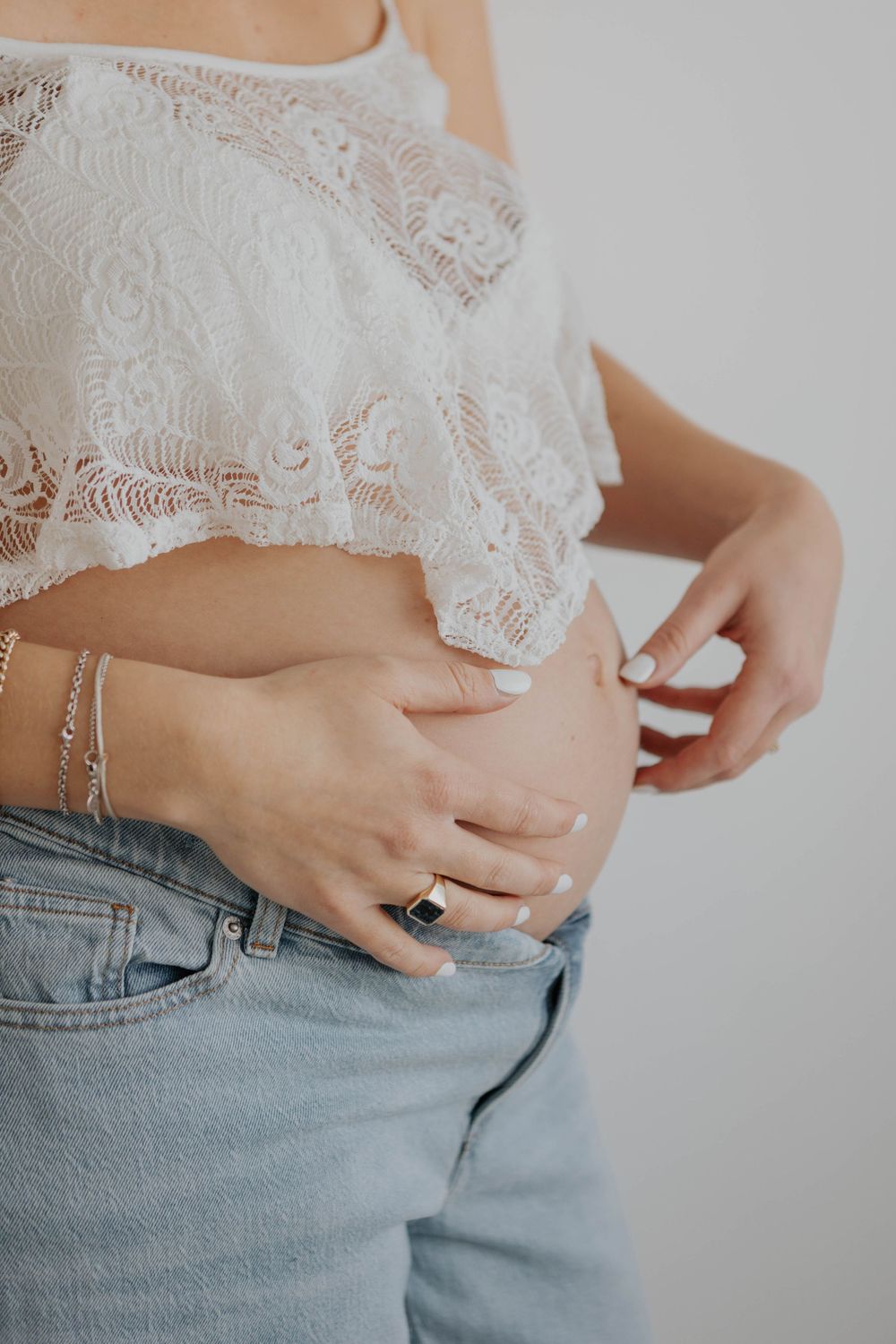 Close-up of pregnant woman in white lace crop top and jeans touching bare belly. Cornwall Maternity Photographer