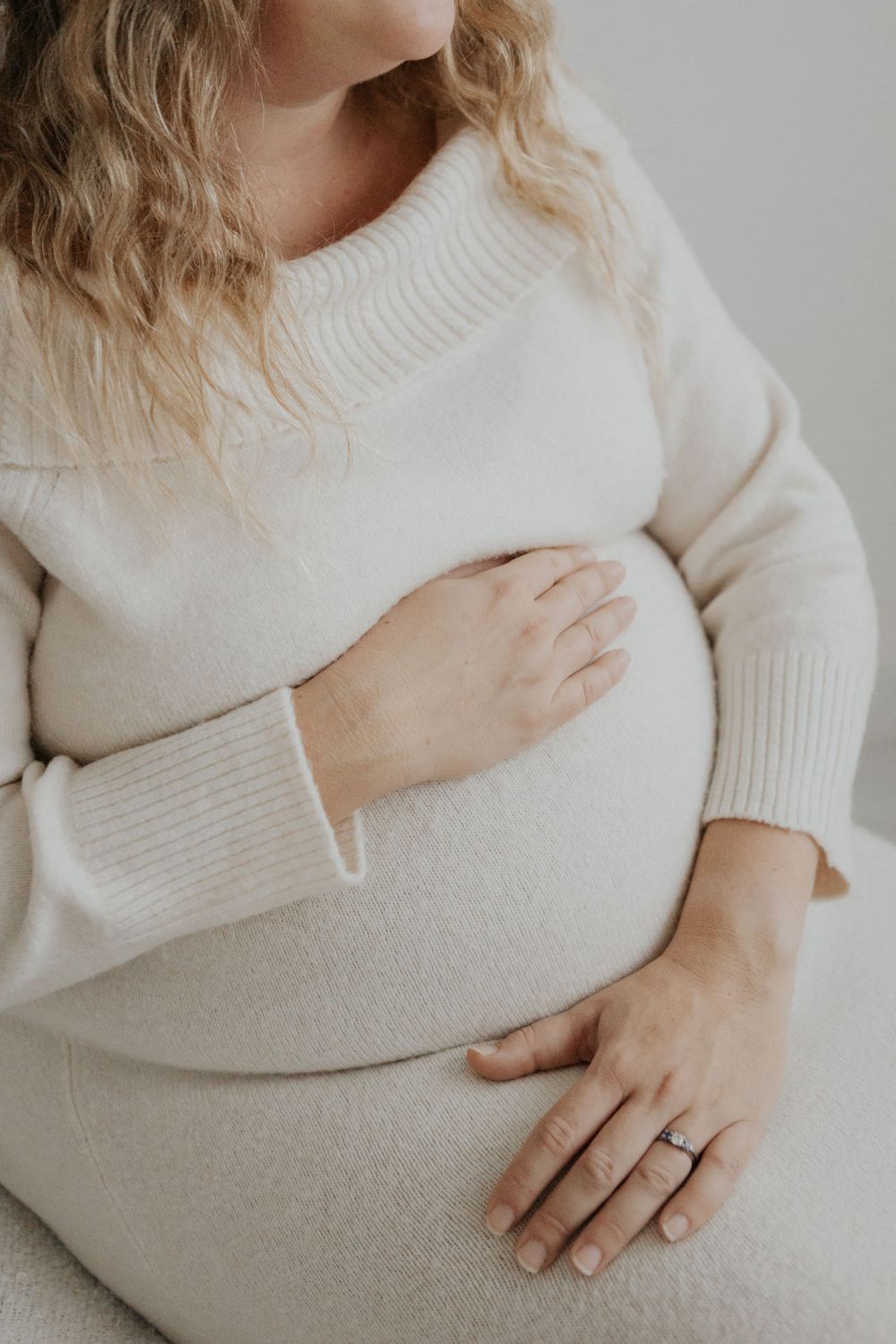 Pregnant woman in cream knit sweater seated and holding baby bump with both hands. Cornwall Maternity Photographer
