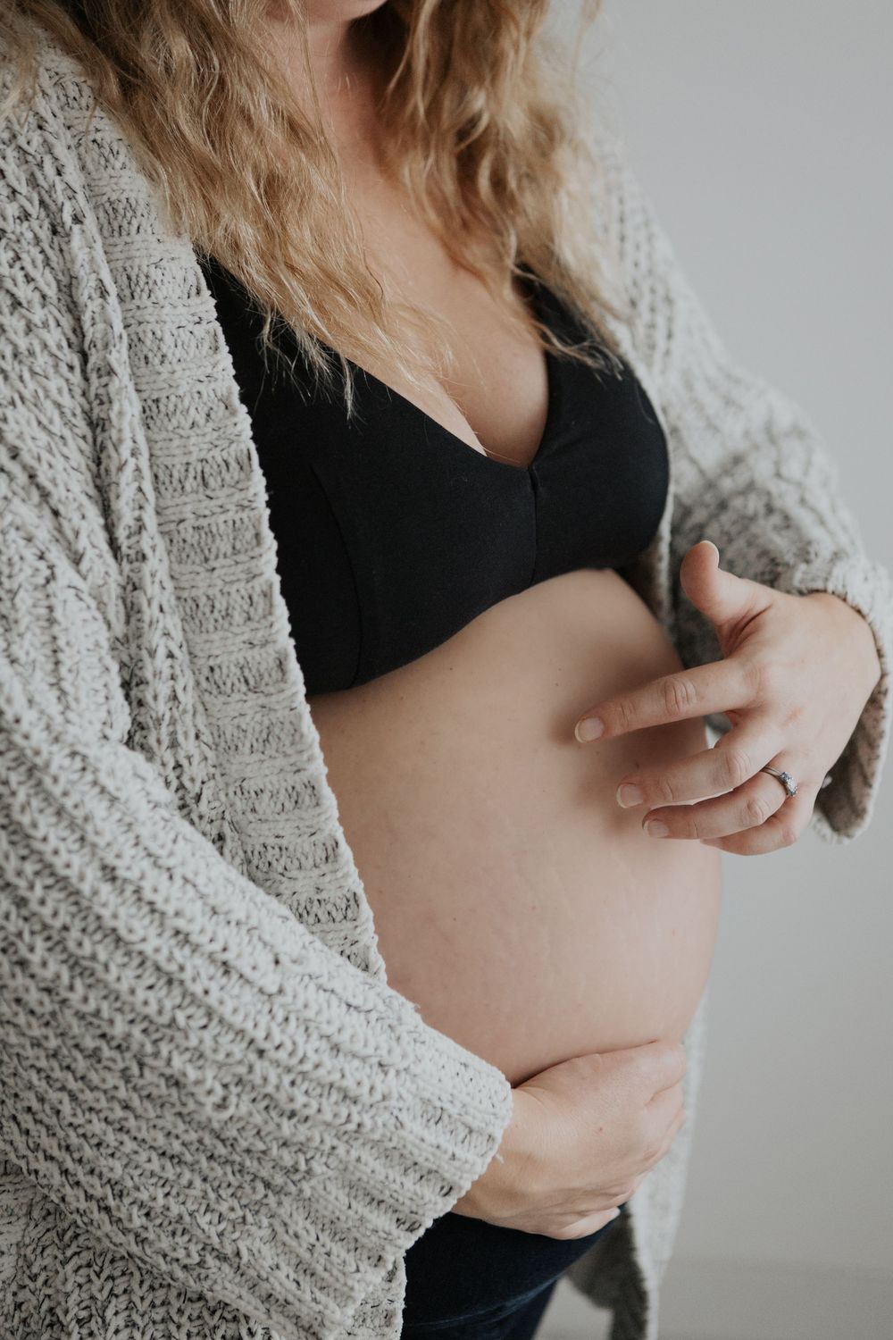 Pregnant woman in black bralette and gray knit cardigan cradling her baby bump with both hands. Cornwall Maternity Photographer