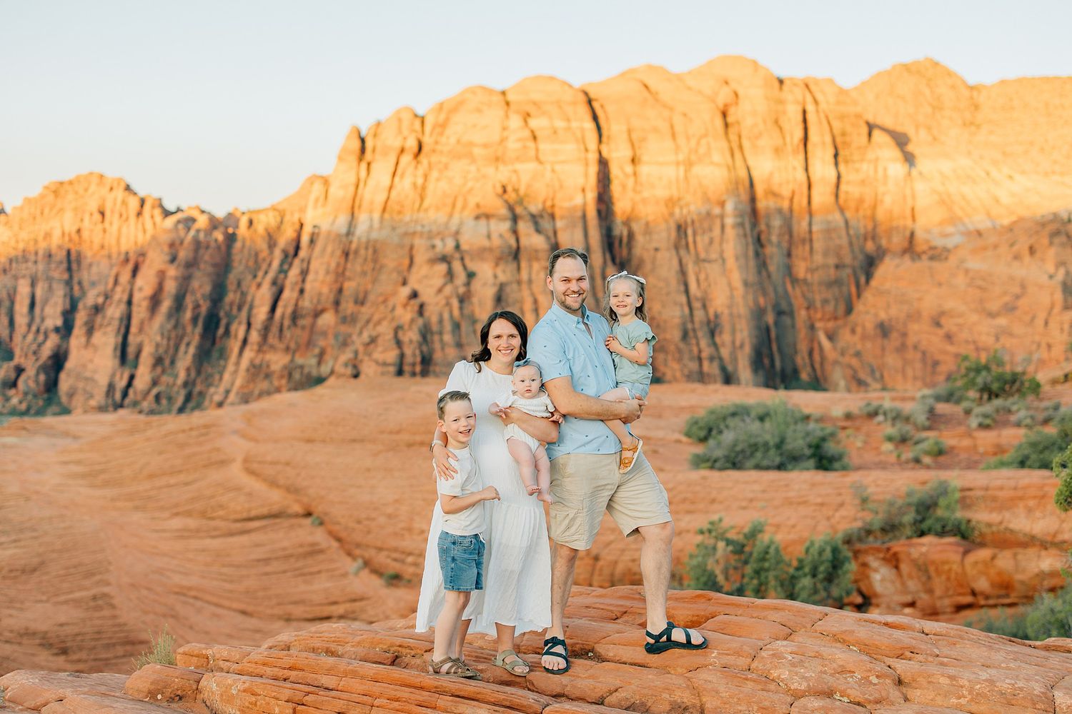 Family of five posing on red rock formations with stunning sandstone cliffs glowing at sunset in the background.