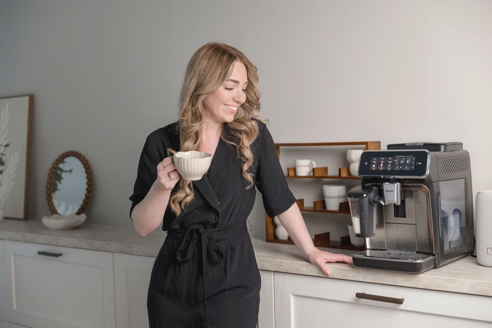 Woman in black dress holding a white coffee cup smiling beside a modern espresso machine in a bright kitchen.