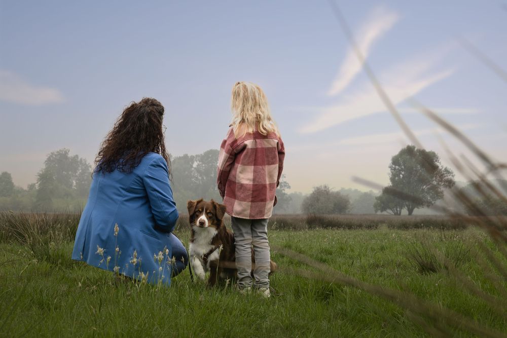 Woman and girl with border collie dog sitting in misty green field, viewed from behind on a calm morning.