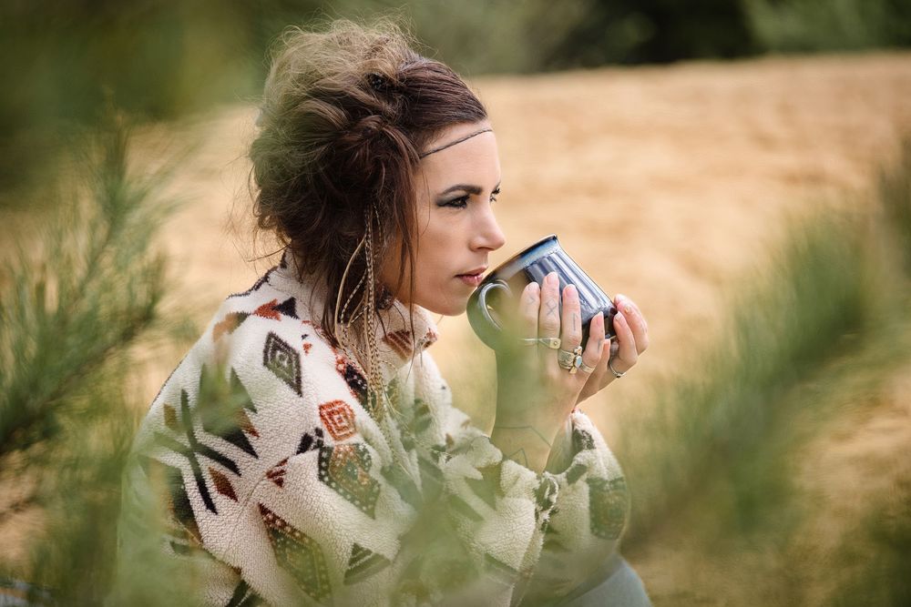 Woman in patterned blanket wrap sipping from a blue enamel mug outdoors in a natural setting.