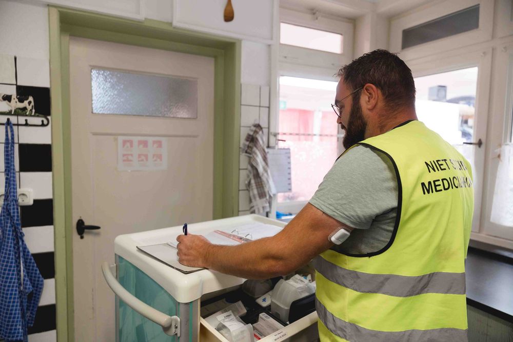A man in a safety vest writes on documents at a medical cart in a clinical room.