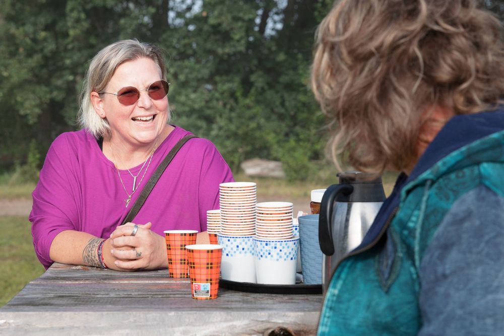 Two women laughing and chatting at a picnic table outdoors with coffee cups and a thermos nearby.