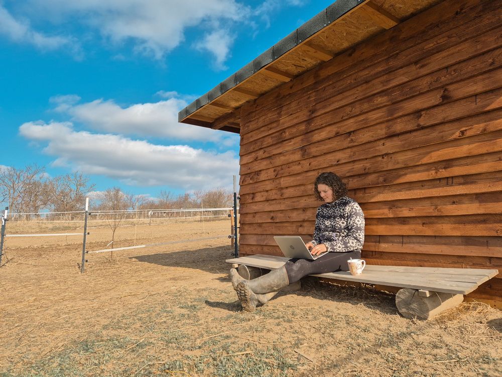 Woman working on laptop outside rustic wooden cabin in rural countryside under blue sky with white clouds.