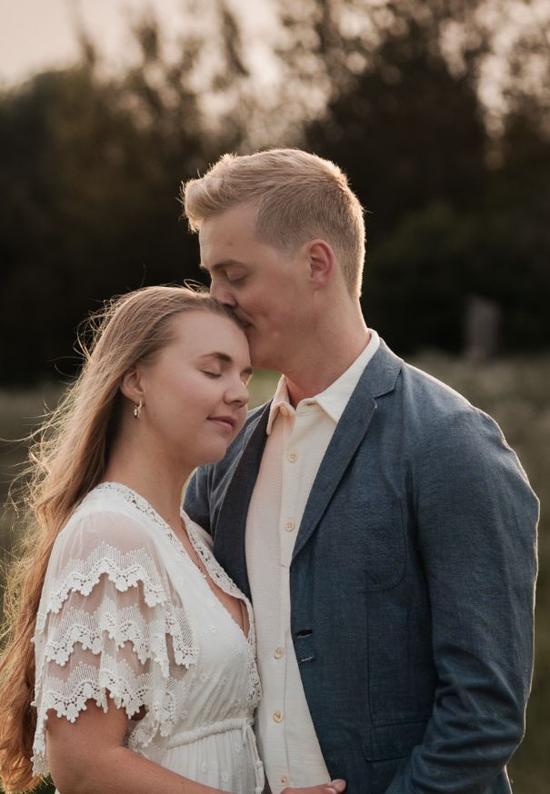 ett par står tillsammans, mannen pussar kvinnan på huvudet, kvinnan ler. kvinnan har en vit klänning och mannen en vit skjorta och blå kavaj