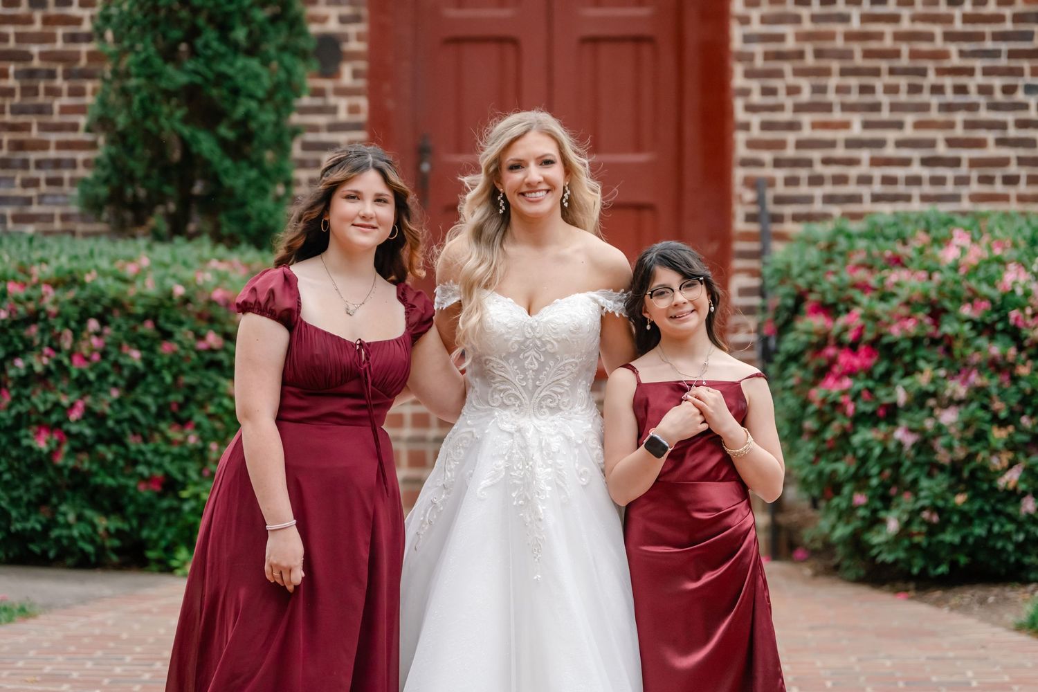 Bride in white lace gown poses with two bridesmaids in burgundy satin dresses outside a brick building.