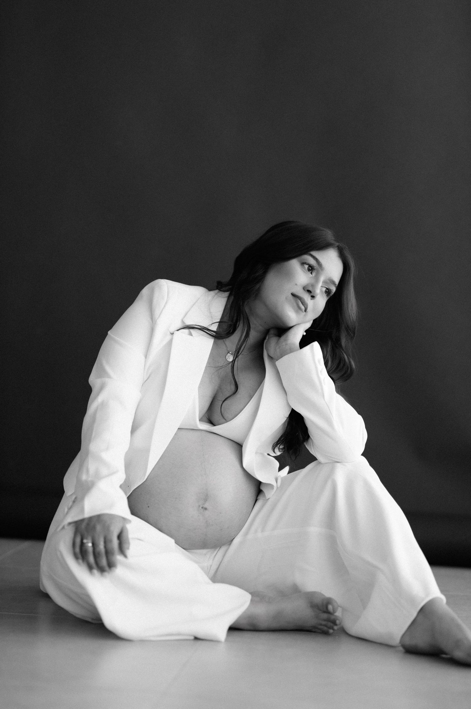Black and white maternity portrait featuring a woman in a white suit jacket and pants reclining on a floor.