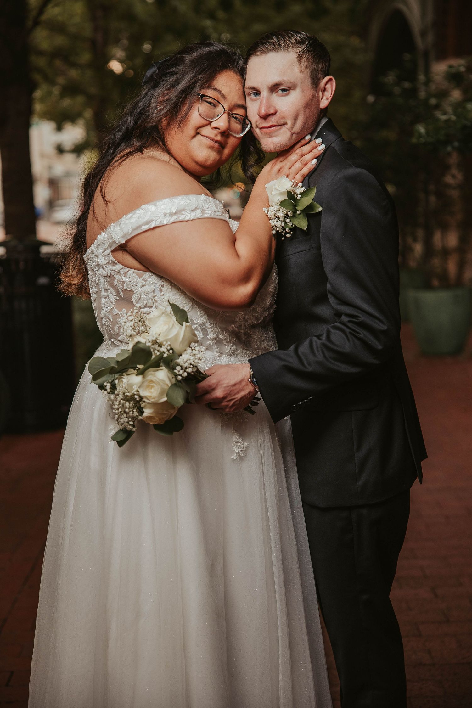 A bride in an off-shoulder lace gown holding white flowers embraces a groom in a dark suit outdoors.