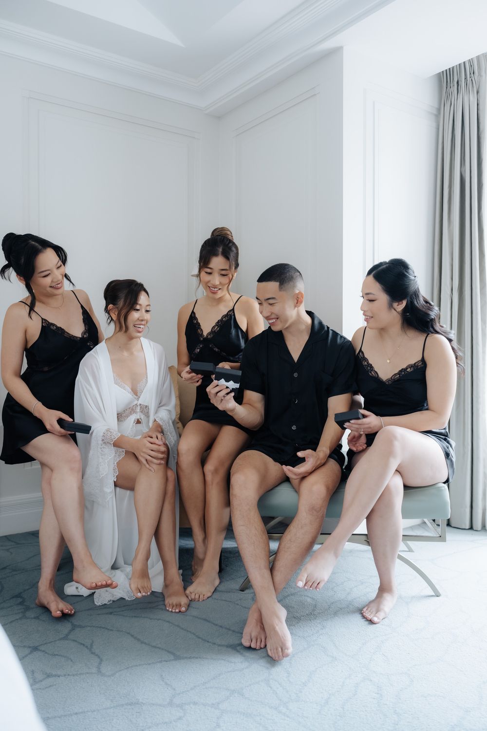 A group of five friends in matching black loungewear sitting together on a bed in a bright hotel room.