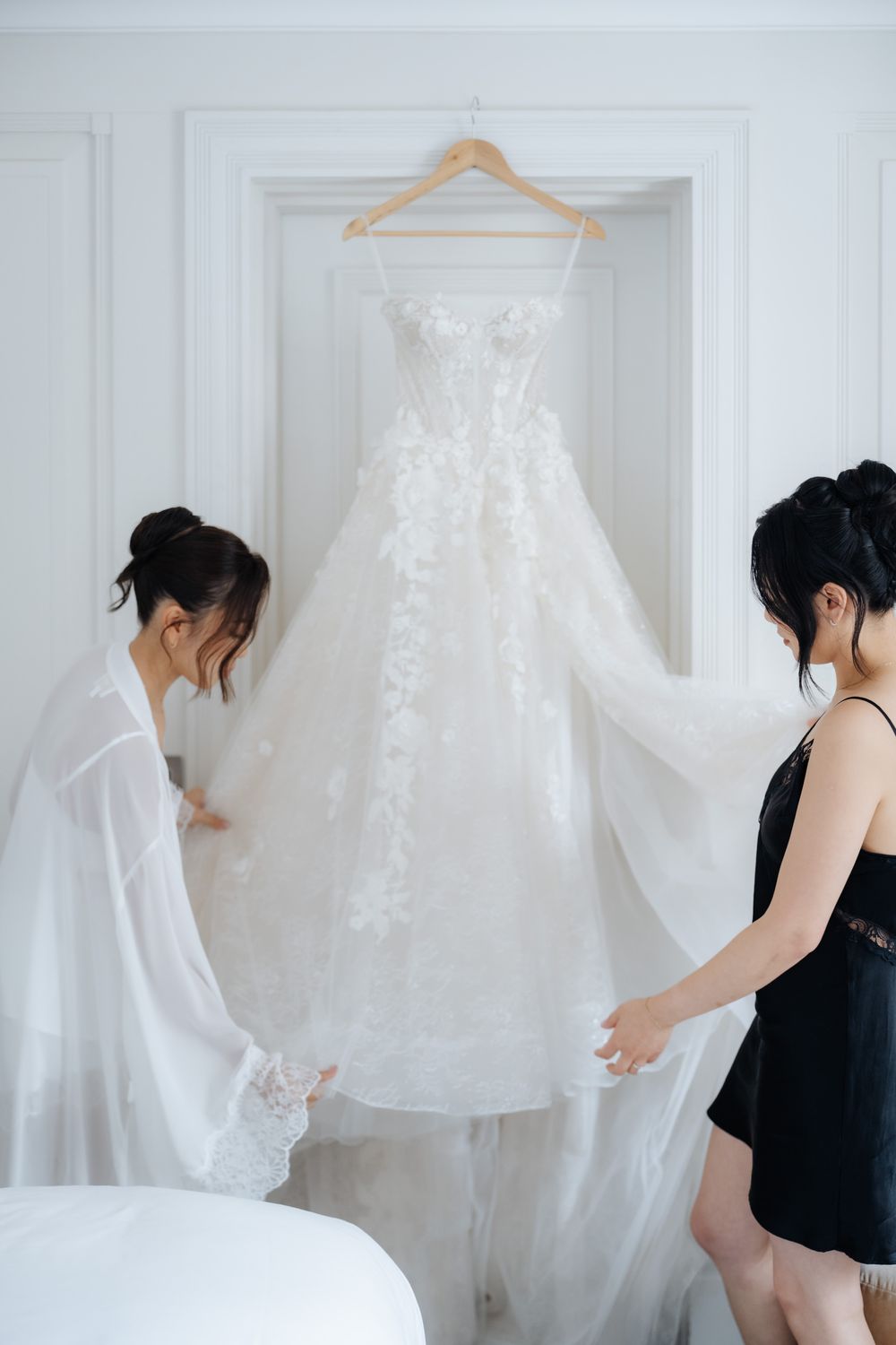 Two women admire a white floral lace wedding gown hanging in a bright white room.