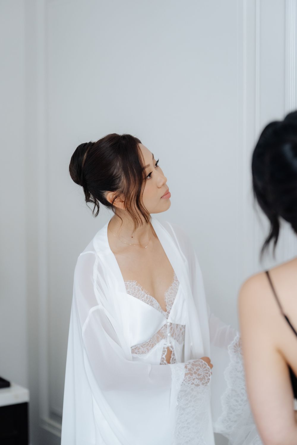Bride in white lace robe and bridal lingerie during wedding morning preparations, elegant updo hairstyle.