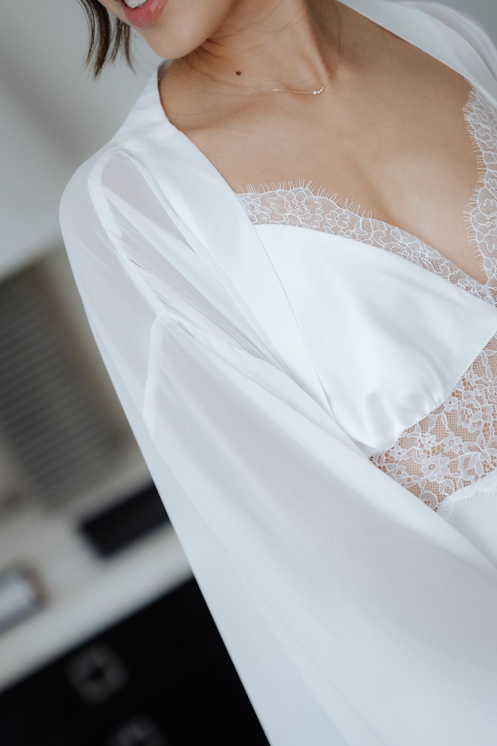 Bride wearing white lace lingerie and sheer robe with delicate gold necklace during wedding morning preparation.