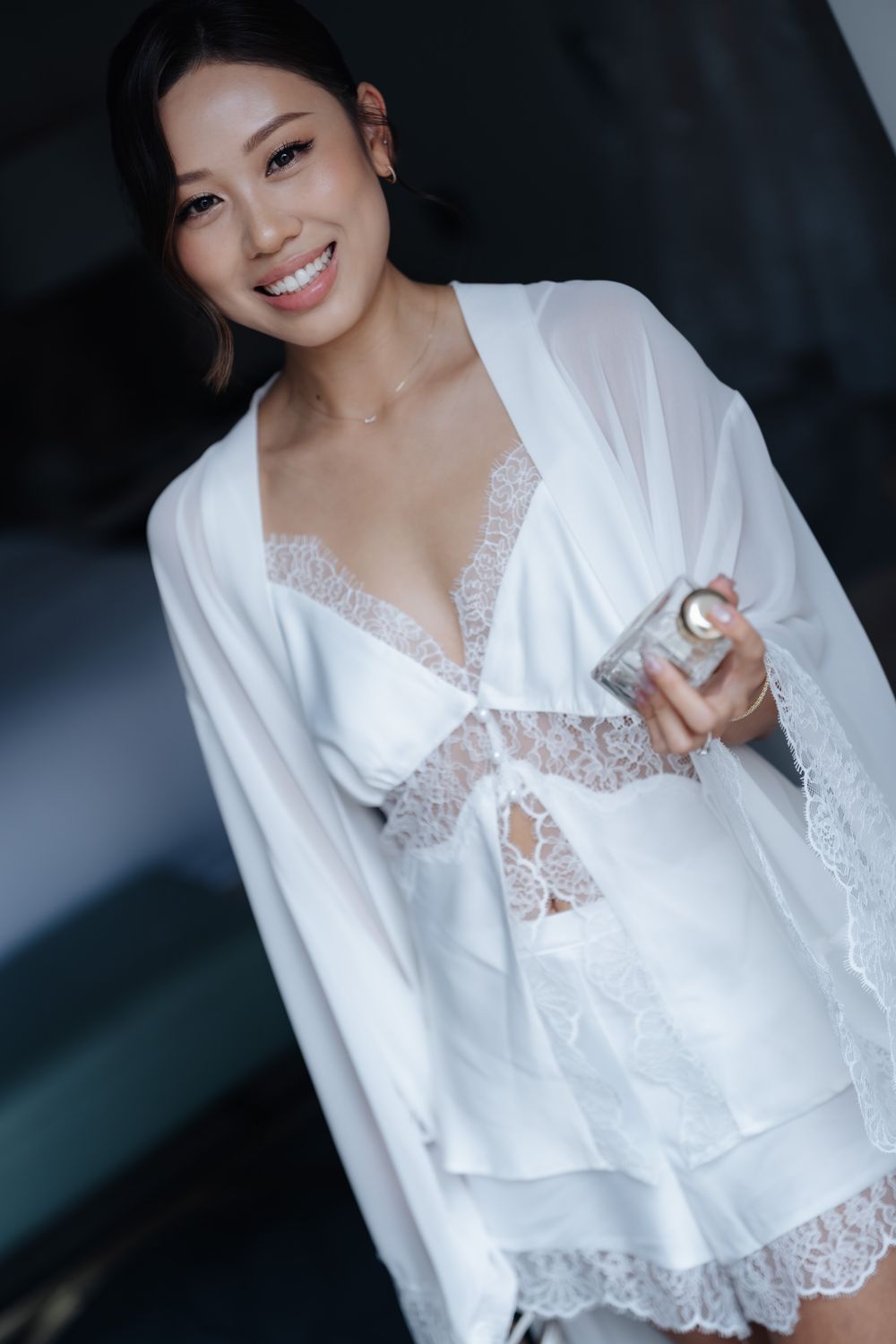 Smiling woman in white lace lingerie set and sheer robe holding a perfume bottle.
