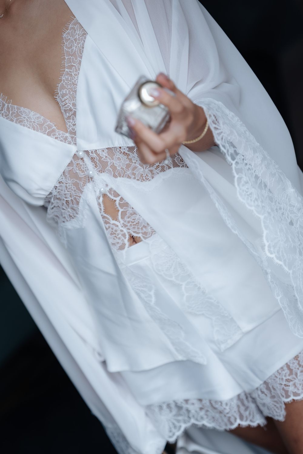 Woman in white lace bridal robe holding a perfume bottle, elegant wedding morning preparation.