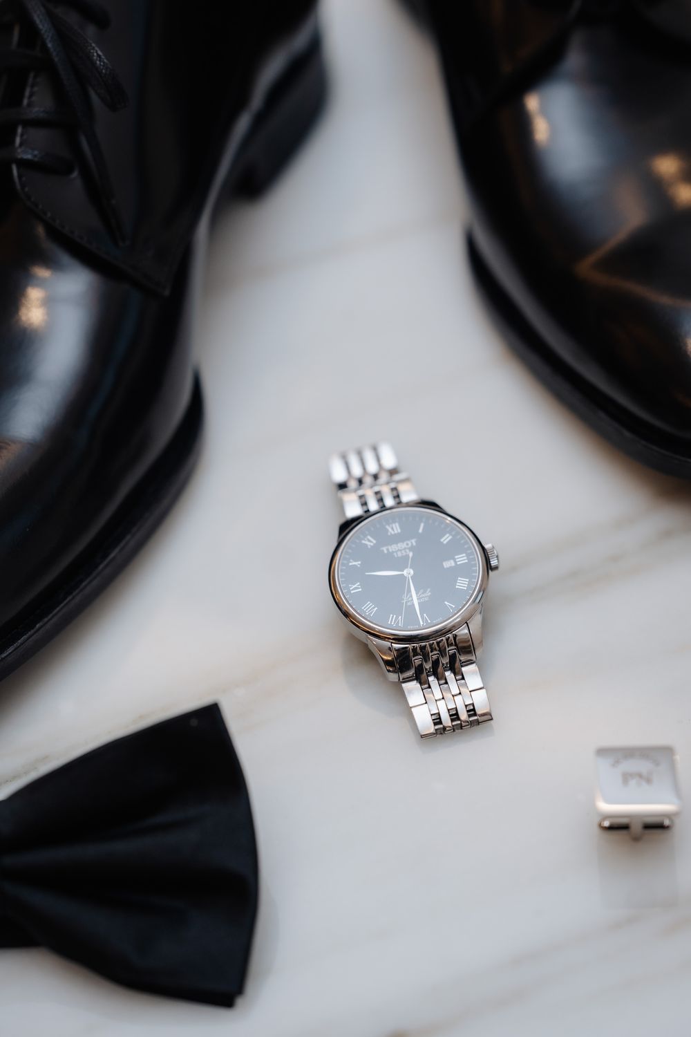 Silver watch with black dial, black bow tie, cufflinks, and polished black dress shoes on marble surface.