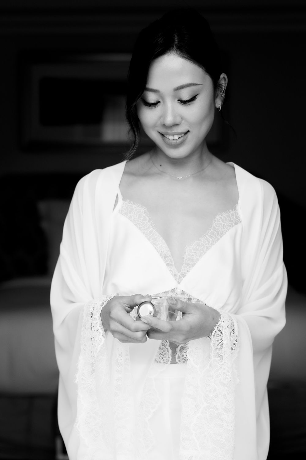 Smiling bride in white lace robe holding a small keepsake item while getting ready on her wedding day.