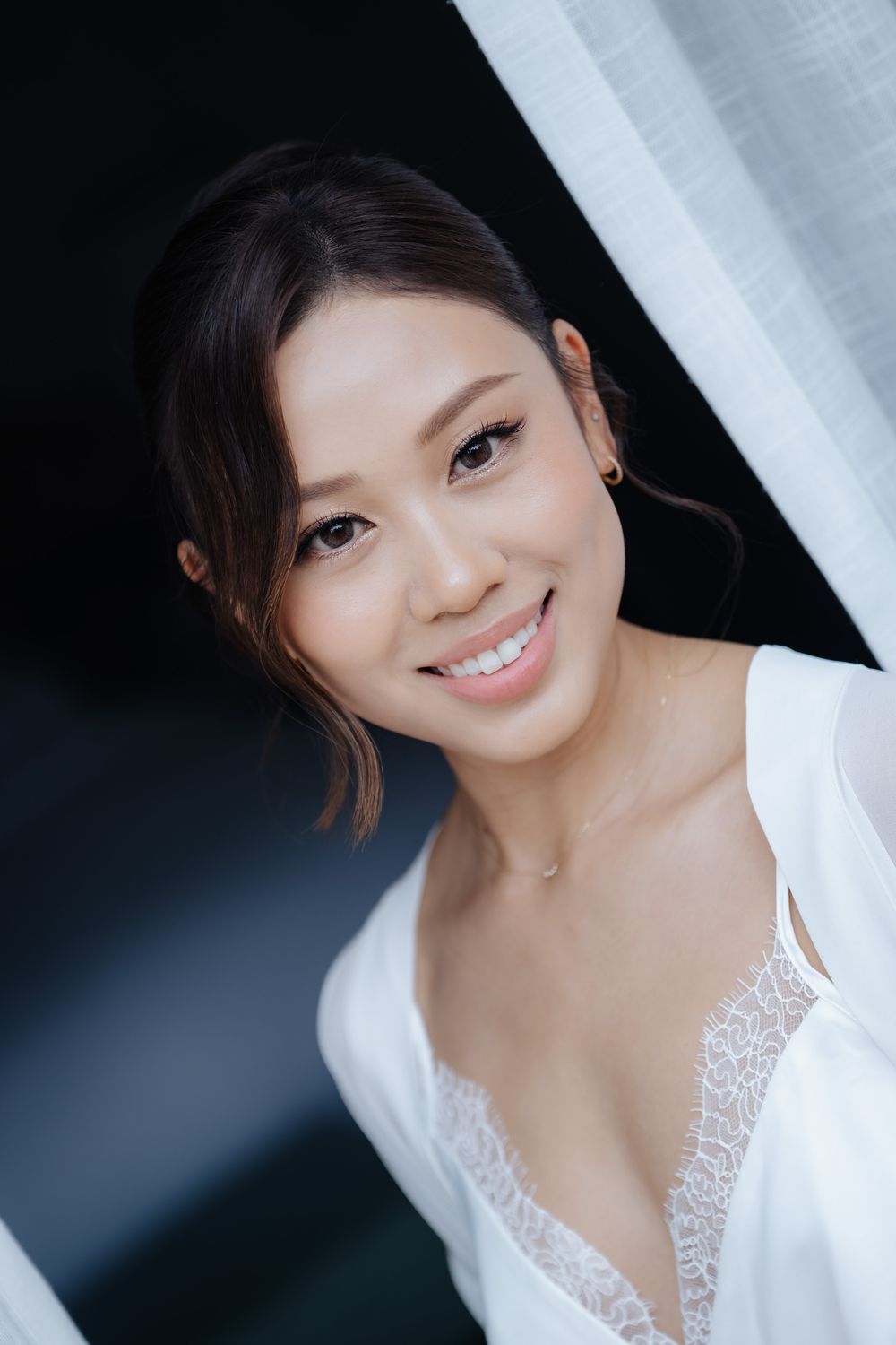 Smiling young Asian woman in white lace top poses near sheer curtain with dark background.