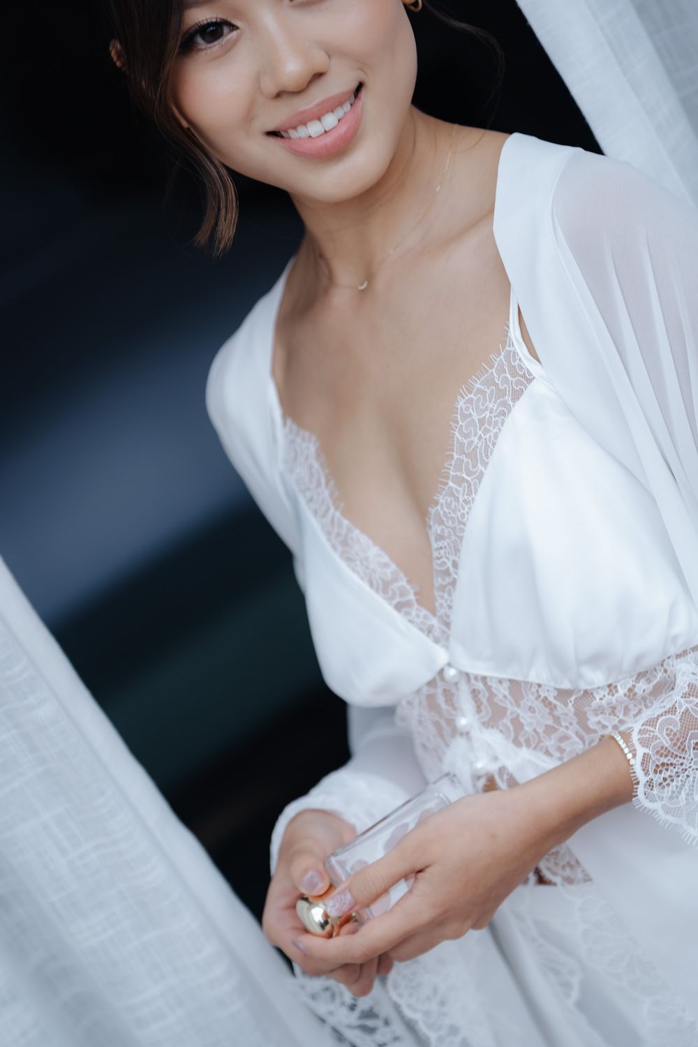 Smiling bride in white lace robe holding perfume bottle, wearing veil and delicate necklace on her wedding day.