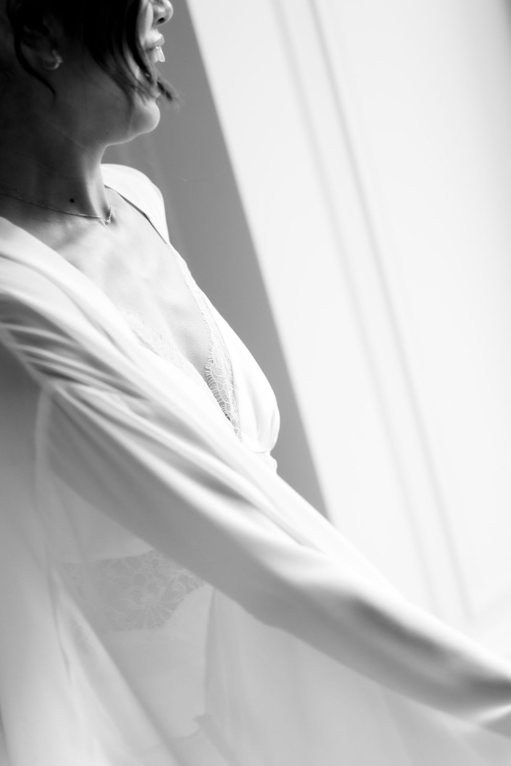 Black and white photo of a bride in a white robe standing by a window on her wedding morning.