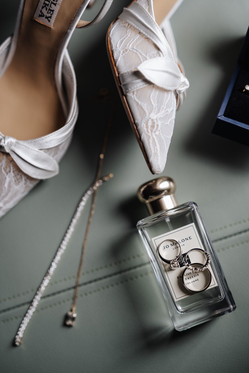 White lace bridal heels, Jo Malone perfume bottle, gold rings, and delicate necklace arranged on sage green surface.