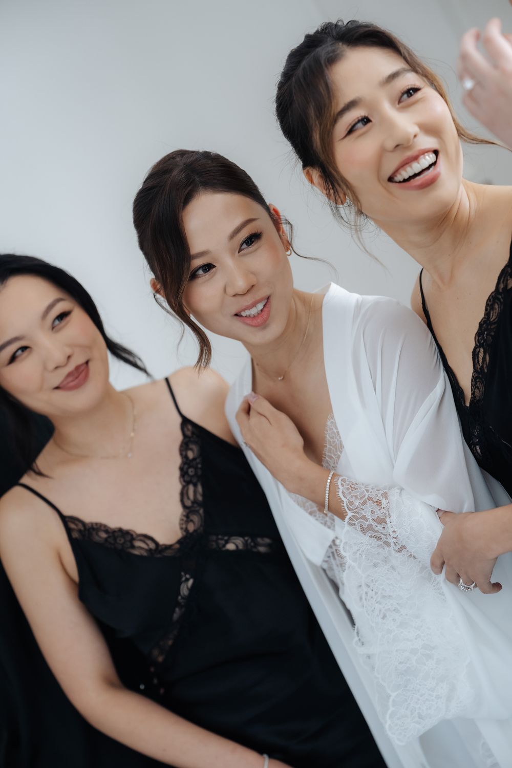 Three smiling Asian women pose together, one in a white bridal robe and two in black lace dresses, celebrating a wedding day.