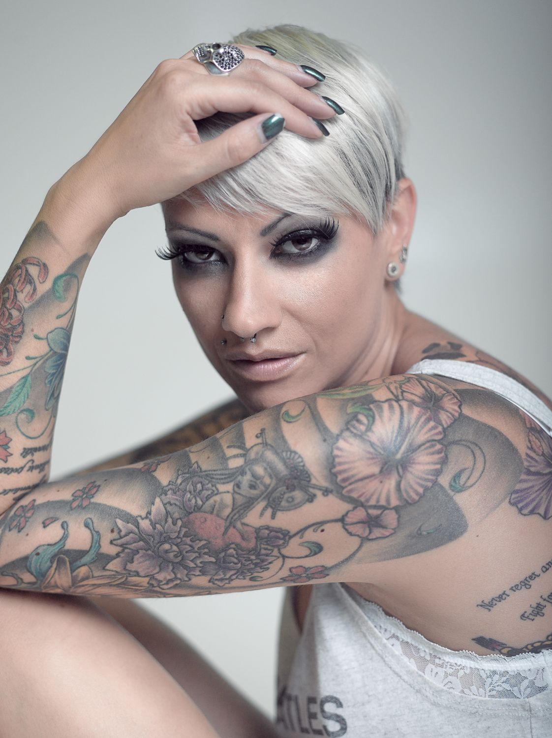 Heavily tattooed blonde woman with short hair and dark makeup posing in white tank top portrait.