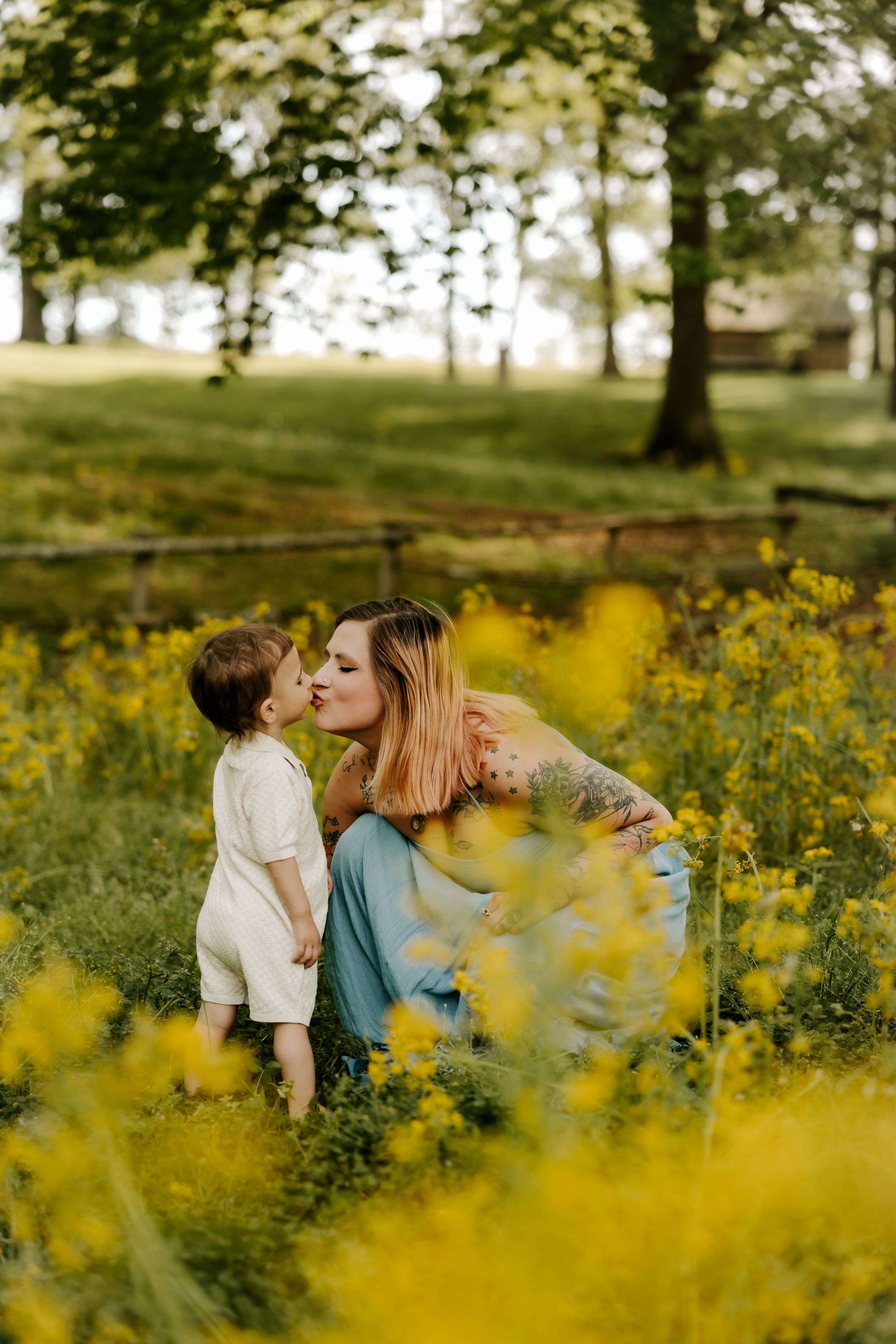 A mother and toddler share a sweet kiss in a vibrant yellow wildflower field with lush green trees in the background.