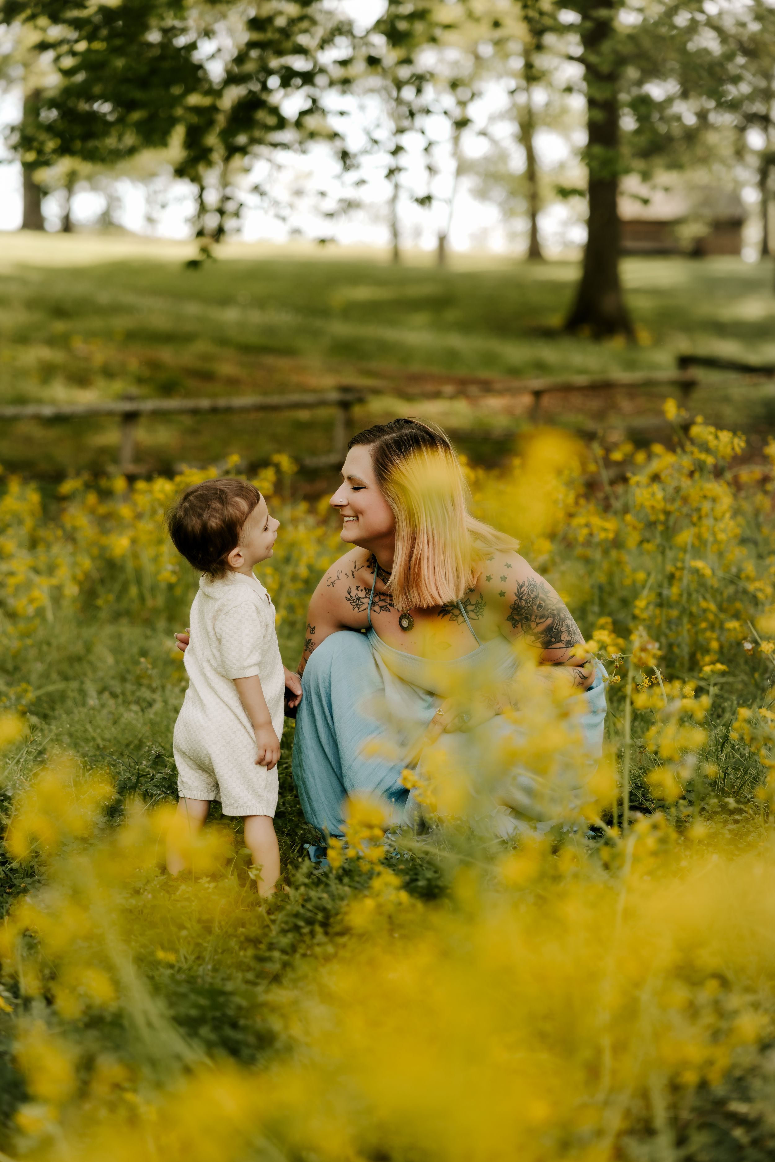 A tattooed mother smiles at her toddler among yellow wildflowers in a lush green field.