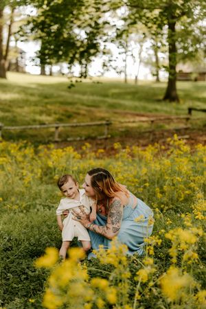 A tattooed mother crouches in a field of yellow wildflowers, lovingly embracing her toddler on a sunny day.