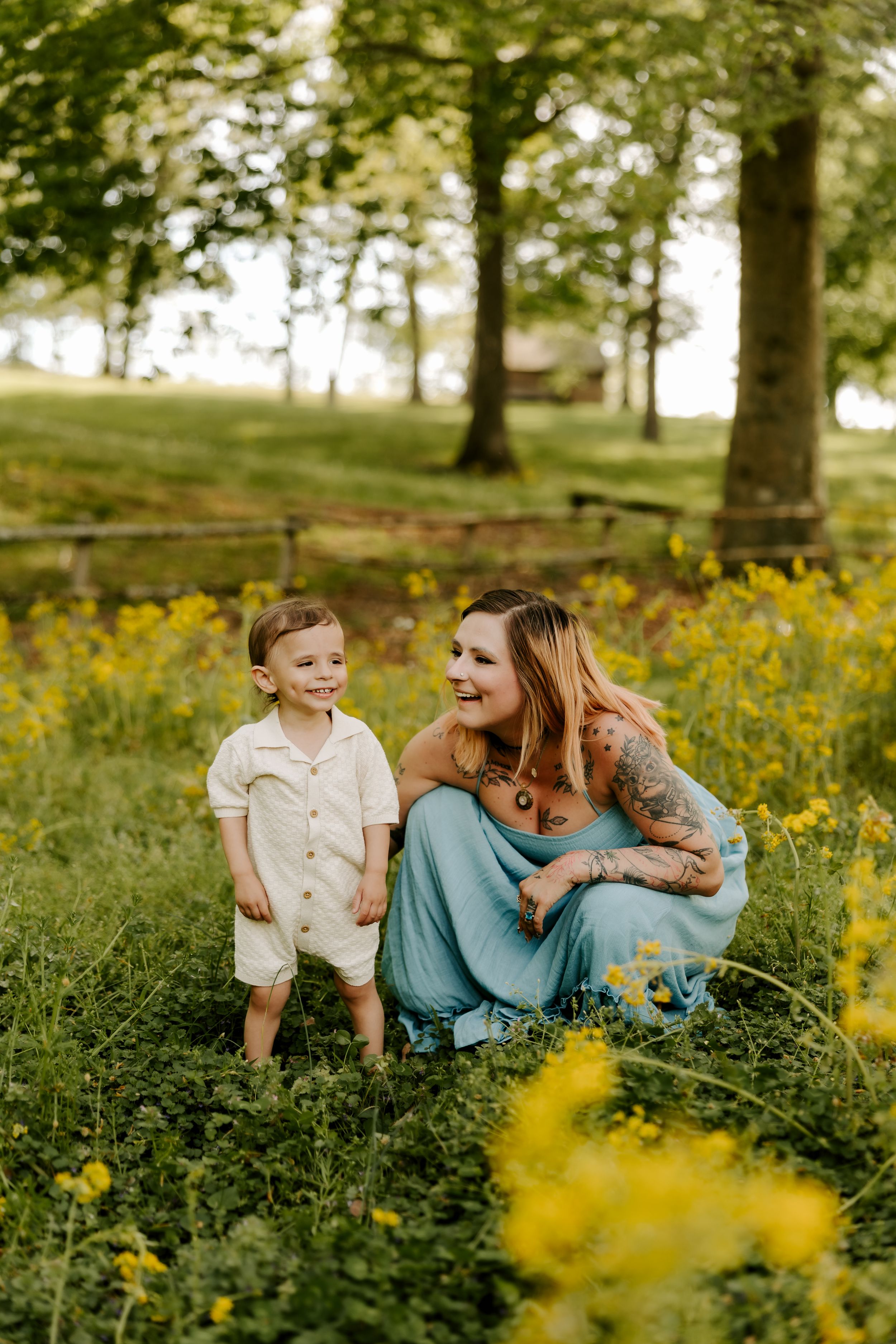 A tattooed mother crouches beside her toddler son in a field of yellow wildflowers, both smiling outdoors.