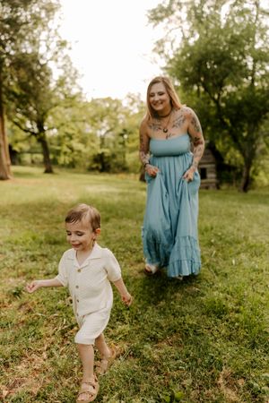 Tattooed mom in blue maxi dress smiles as toddler runs ahead on green grass near rustic cabin.