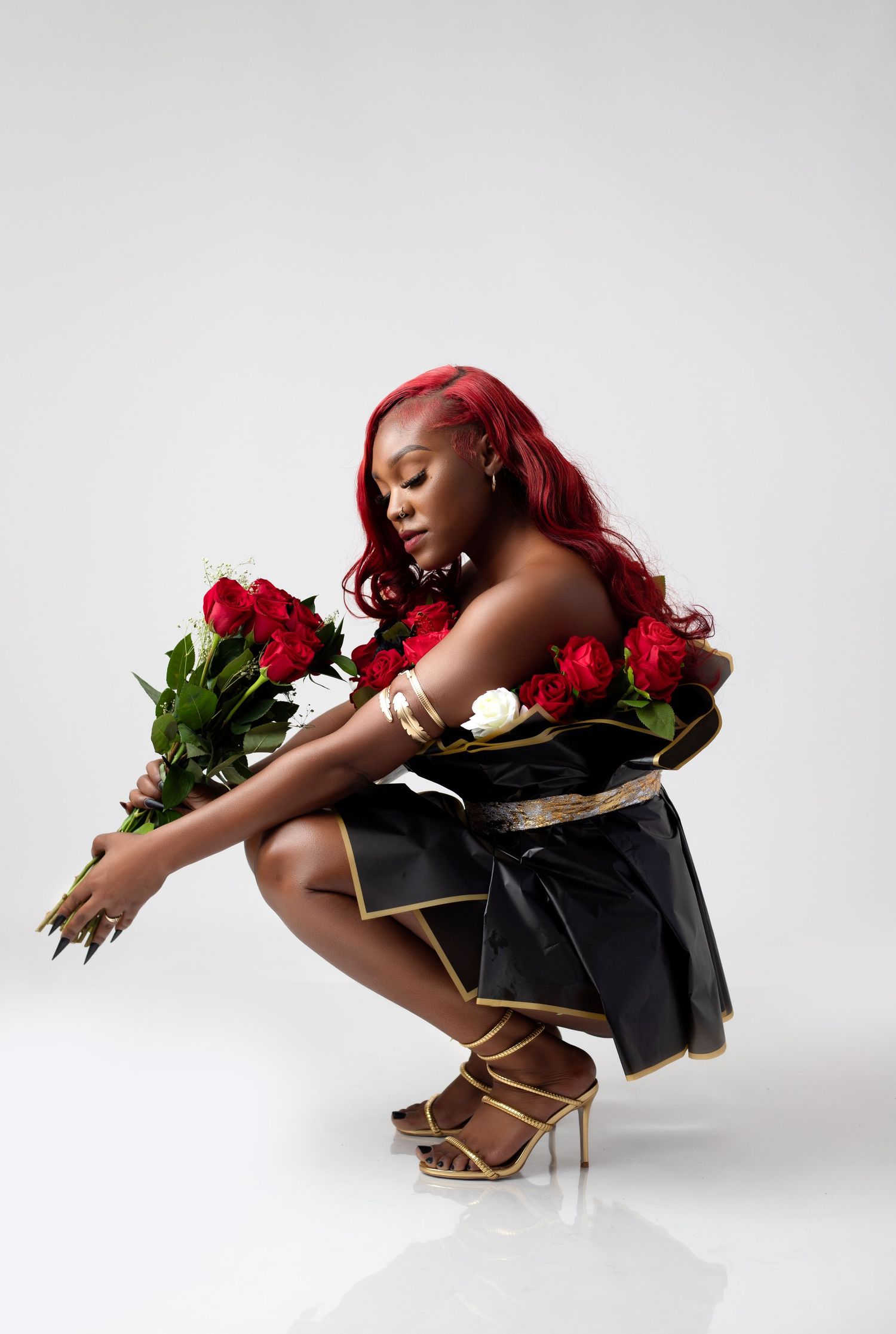 A graceful women in black with vibrant red hair poses with red roses against a white backdrop in a dynamic yet sexy photoshoot.