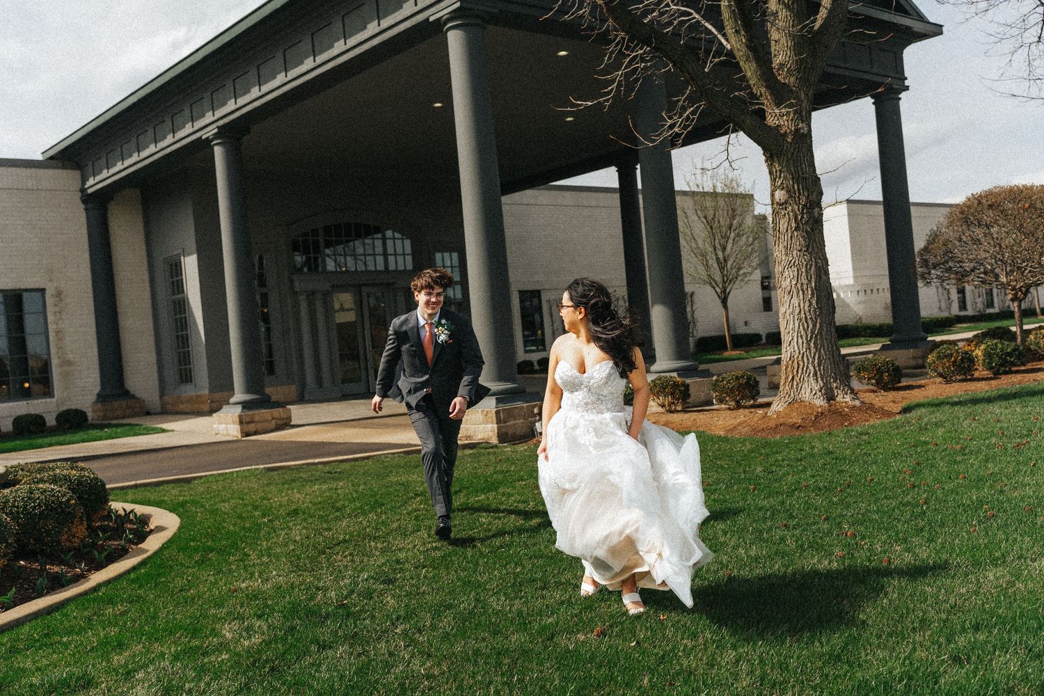 Bride and groom running joyfully across green lawn outside elegant wedding venue with large columns.